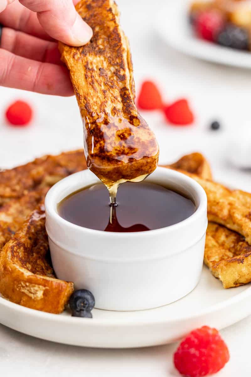 French toast stick being dipped into a bowl of maple syrup.