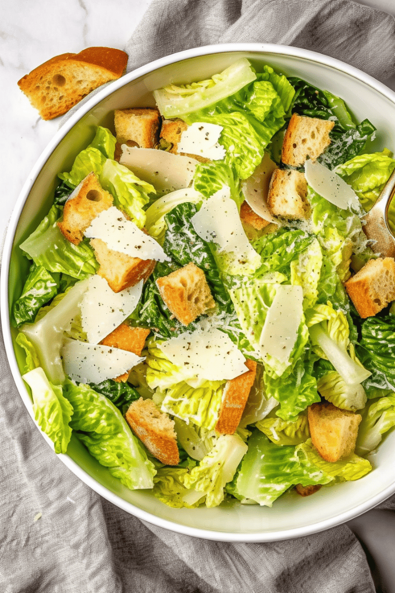 Overhead view of a large bowl of caesar salad with homemade croutons and shaved parmesan on a bed of romaine lettuce.