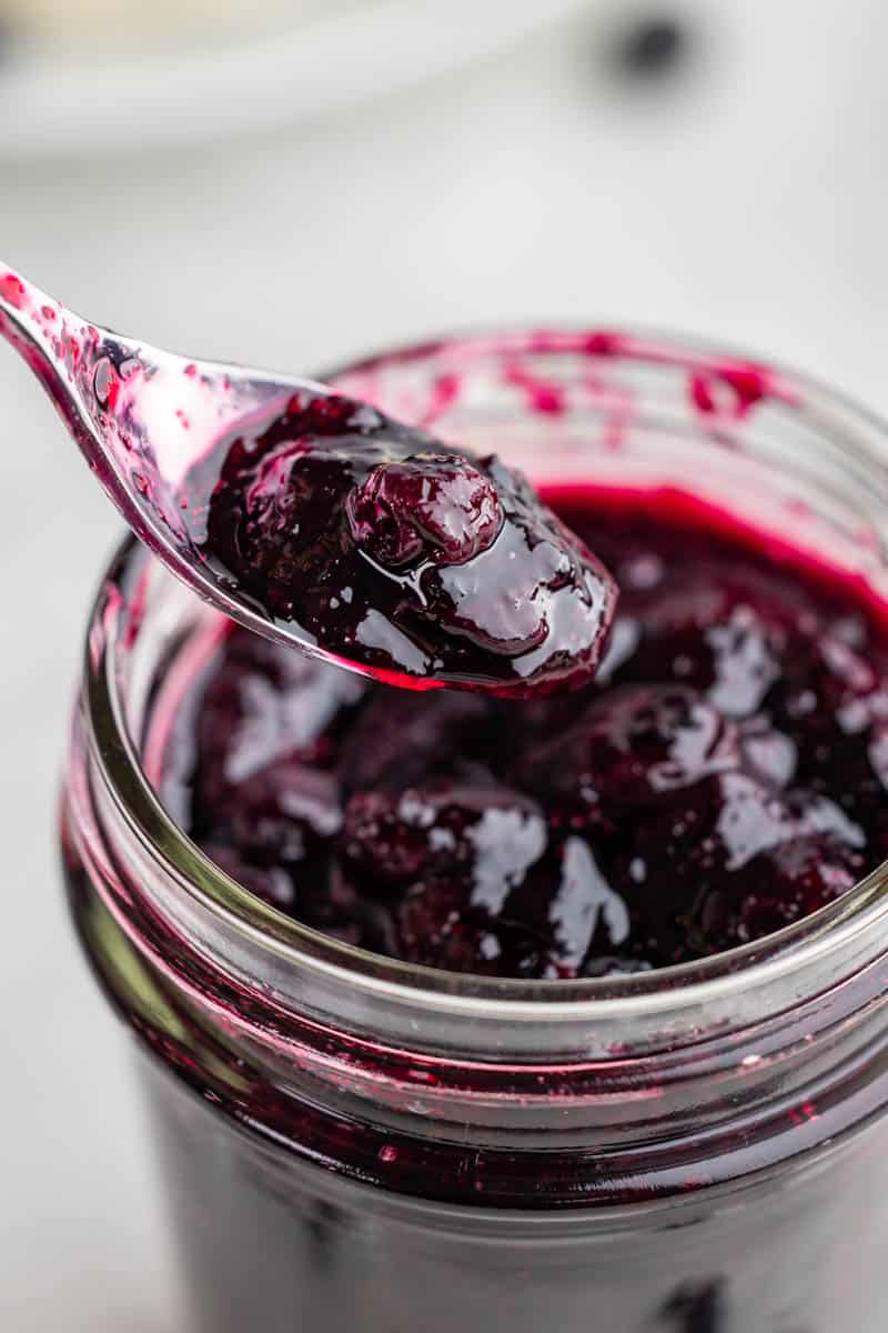 A spoon filled with homemade blueberry syrup held over a glass jar filled with the syrup.
