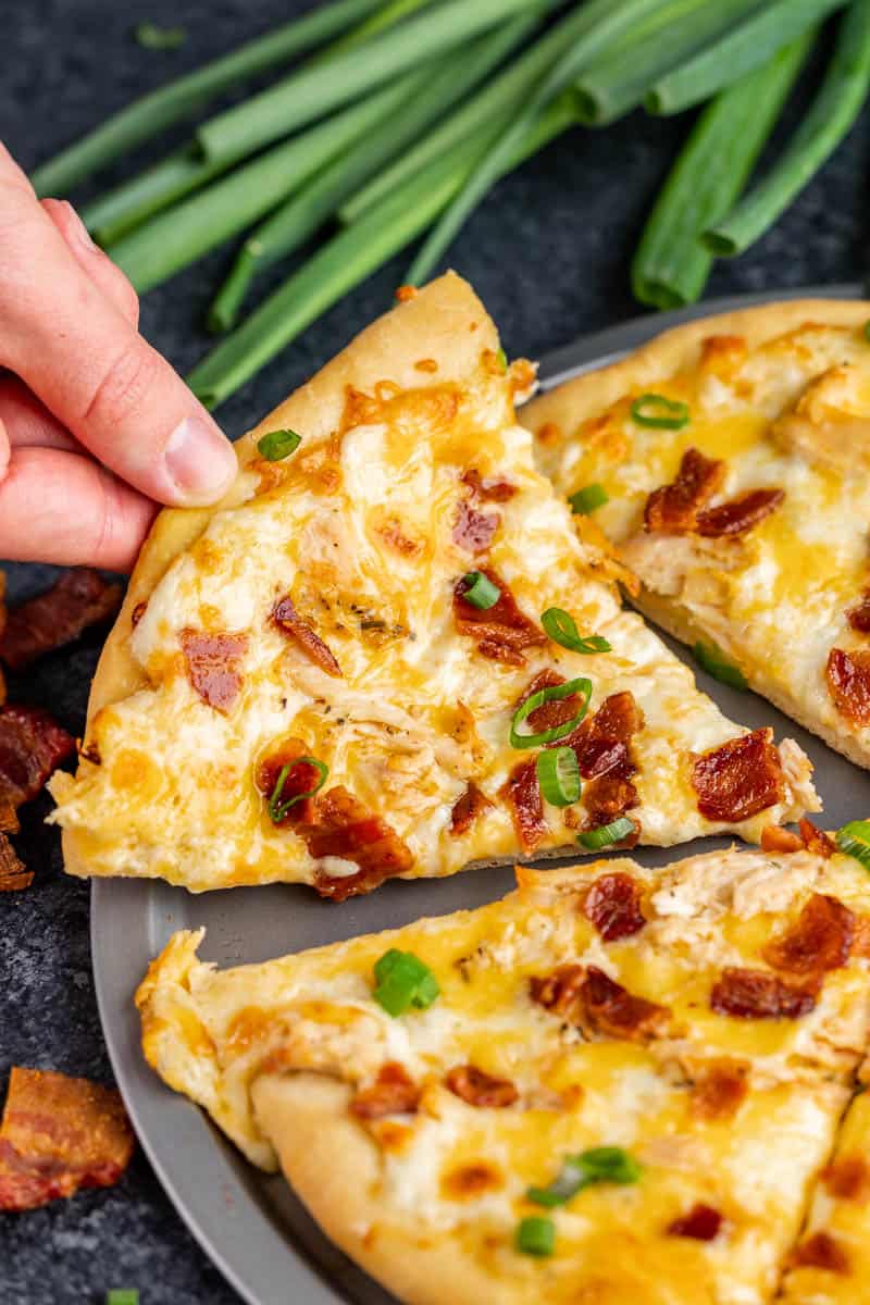 A hand pulling away a slice of chicken bacon ranch pizza from the pan.
