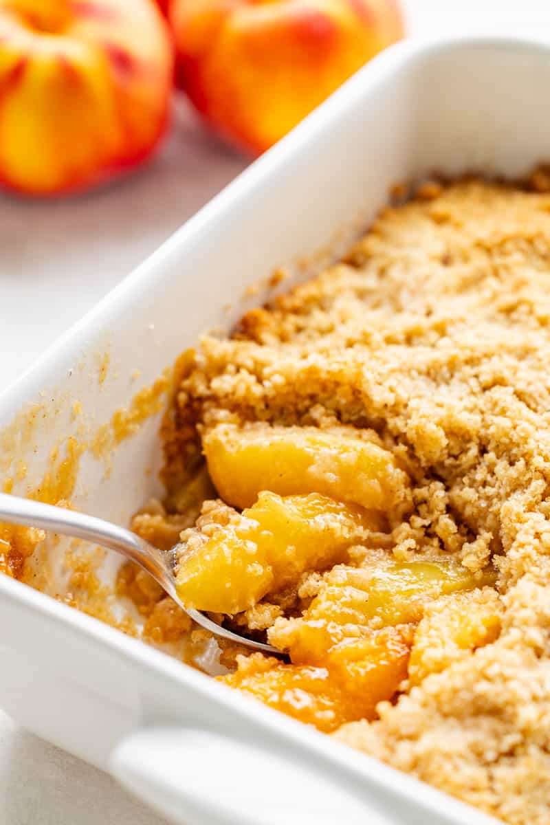 A spoon dipping into peach crumble in a baking pan.