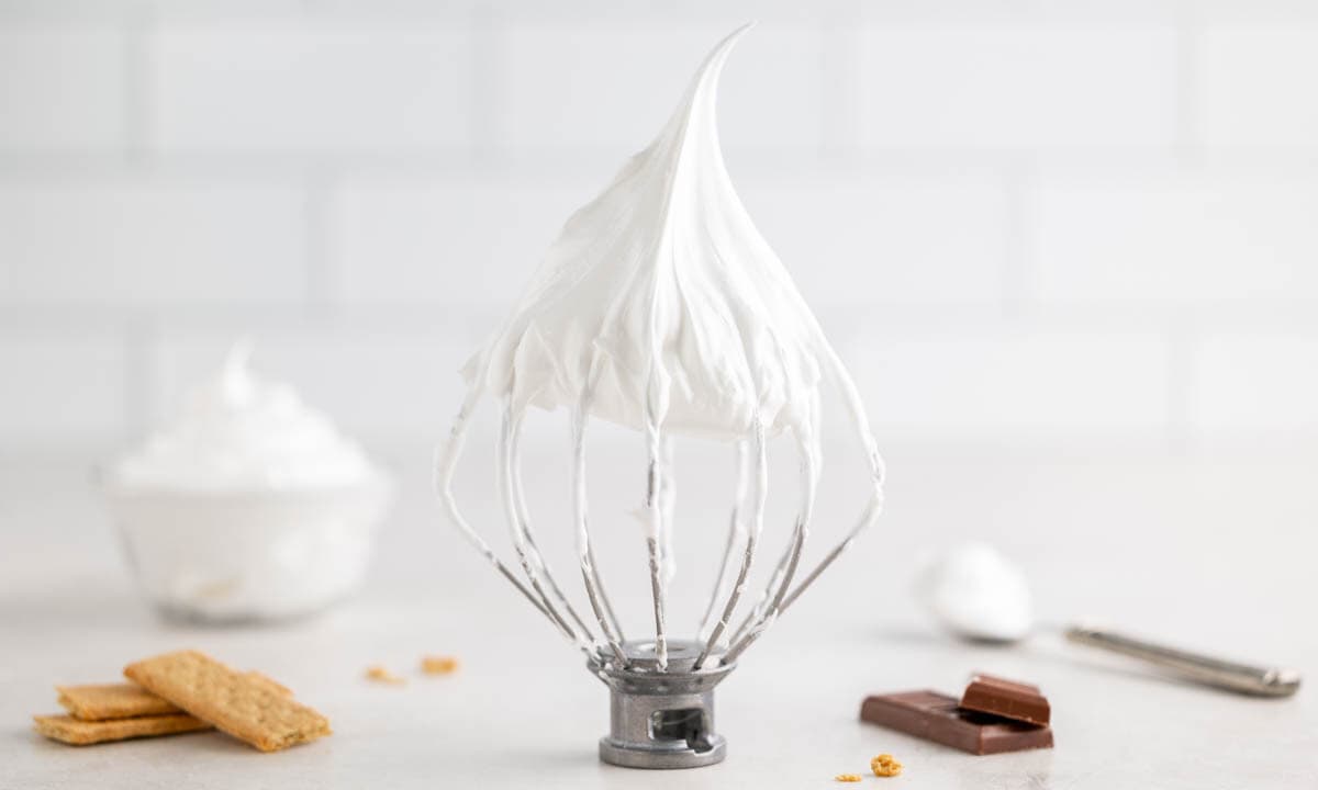 A stand mixer whisk attachment coated in homemade marshmallow fluff, surrounded by graham crackers and chocolate pieces.