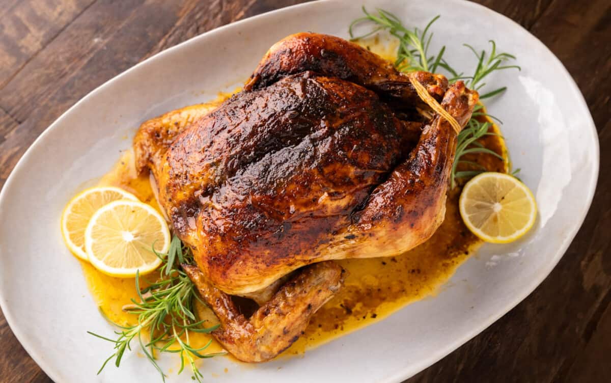A whole, roasted chicken on a whit serving platter with lemon slices and fresh rosemary sprigs.