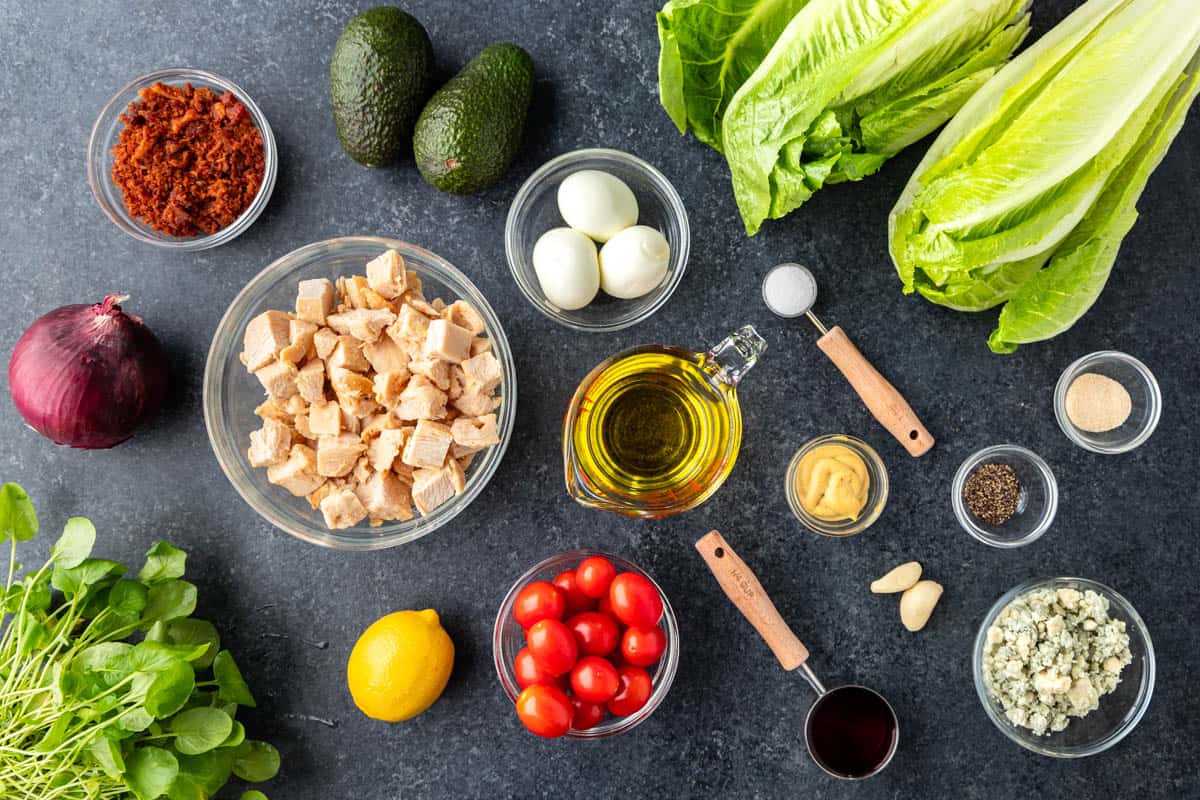 Overhead view of the raw ingredients needed to make Cobb Salad on a clean, dark kitchen counter.