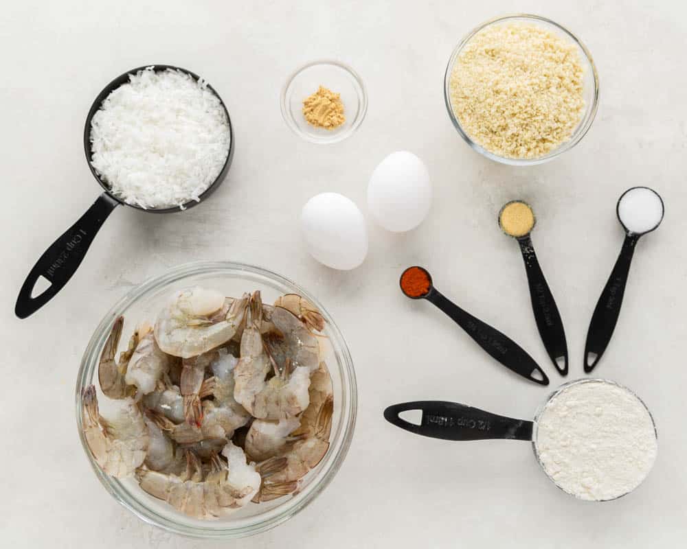 Overhead view of the raw ingredients needed to make crispy coconut shrimp, spread out on a clean kitchen counter.
