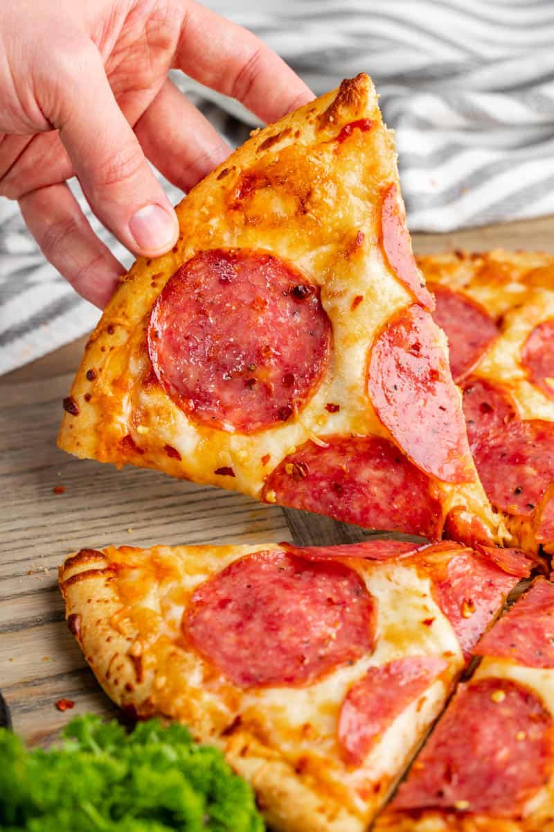 A side view of a hand picking up a slice of salami pizza.