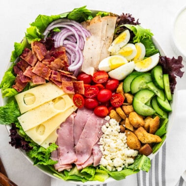 Overhead view of a chef salad with dressing in a small ceramic bowl and wooden serving spoons on the side.