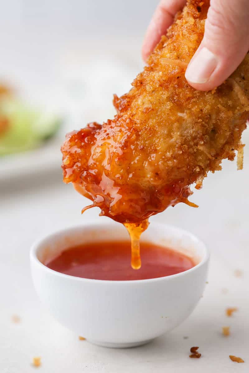 A hand dipping a coconut chicken tender into sweet chili sauce.
