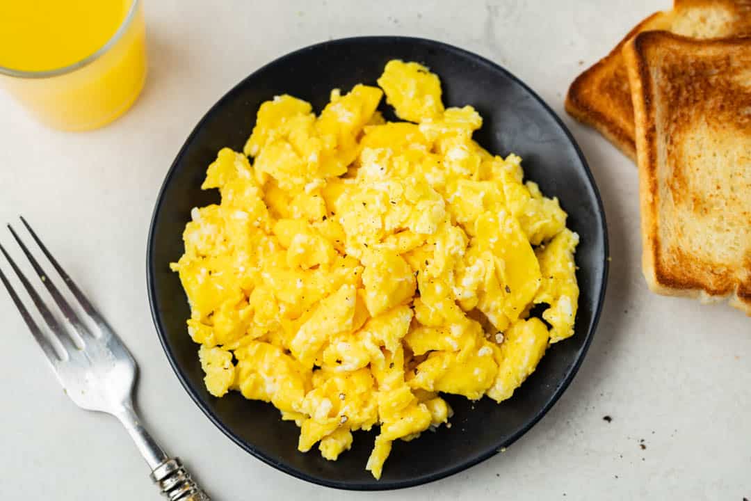 Overhead view of scrambled eggs on a black plate with toast on the side.