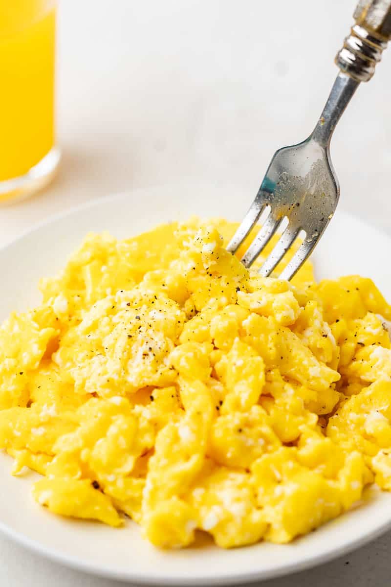 Scrambled eggs with a fork on a white plate with a glass of orange juice in the background.