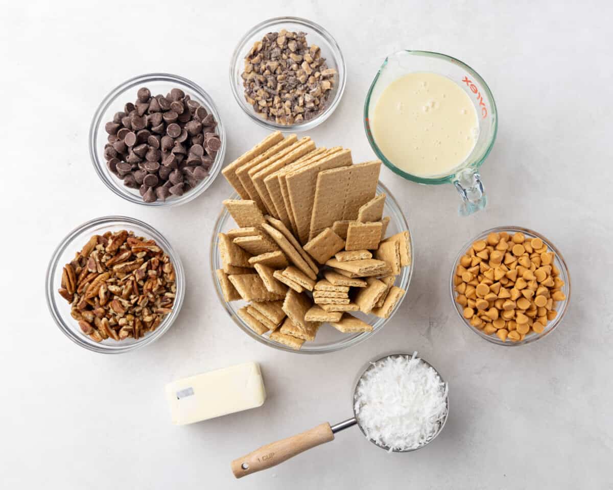 Overhead view of a clean counter with measured out graham crackers, coconut, butter, chocolate chips, sweetened condensed milk, pecans, toffee chips, and peanut butter chips.