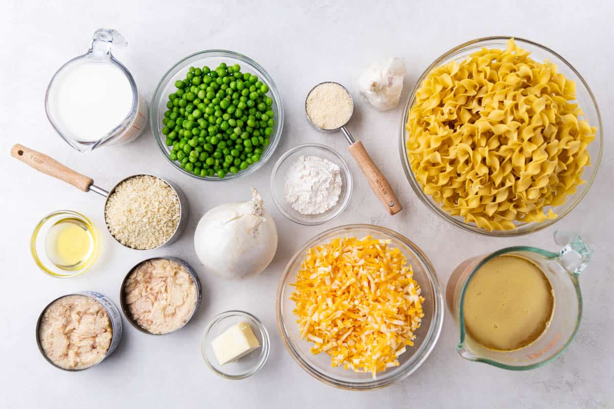 Overhead view of a kitchen countertop with a bowl of noodles, cheese, peas, tuna, breadcrumbs, parmesan, garlic, flour, cream, chicken broth, butter, and an onion.