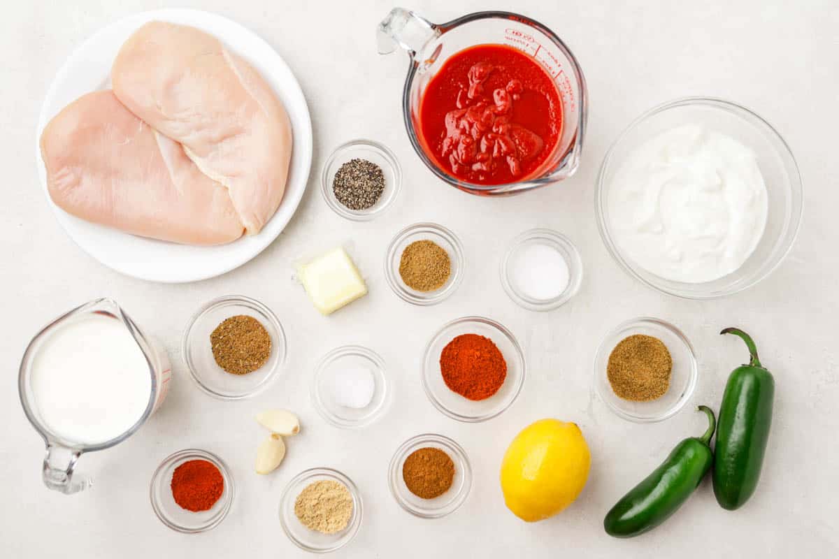 Overhead view of a kitchen counter with measured out spices in individual glass bowls, tomato sauce, cream, garlic, butter, a lemon, jalapeños, and a plate with raw chicken.