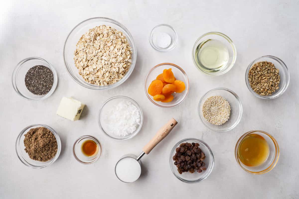 Overhead view of a kitchen counter with bowls of measured out oats, seeds, coconut, dried fruit, butter, and sugar.