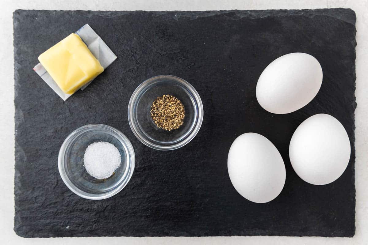 Overhead view of three uncooked eggs, a pat of butter, and small bowls with salt and pepper on a slate slab.
