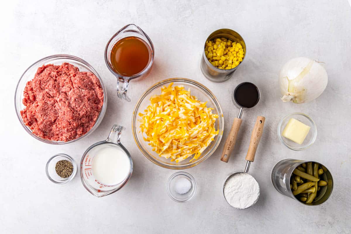 Overhead view of a kitchen counter top with measured out ground beef, broth, milk, salt, pepper, cheese, corn, green beans, butter, onion, flour, and Worcestershire sauce.