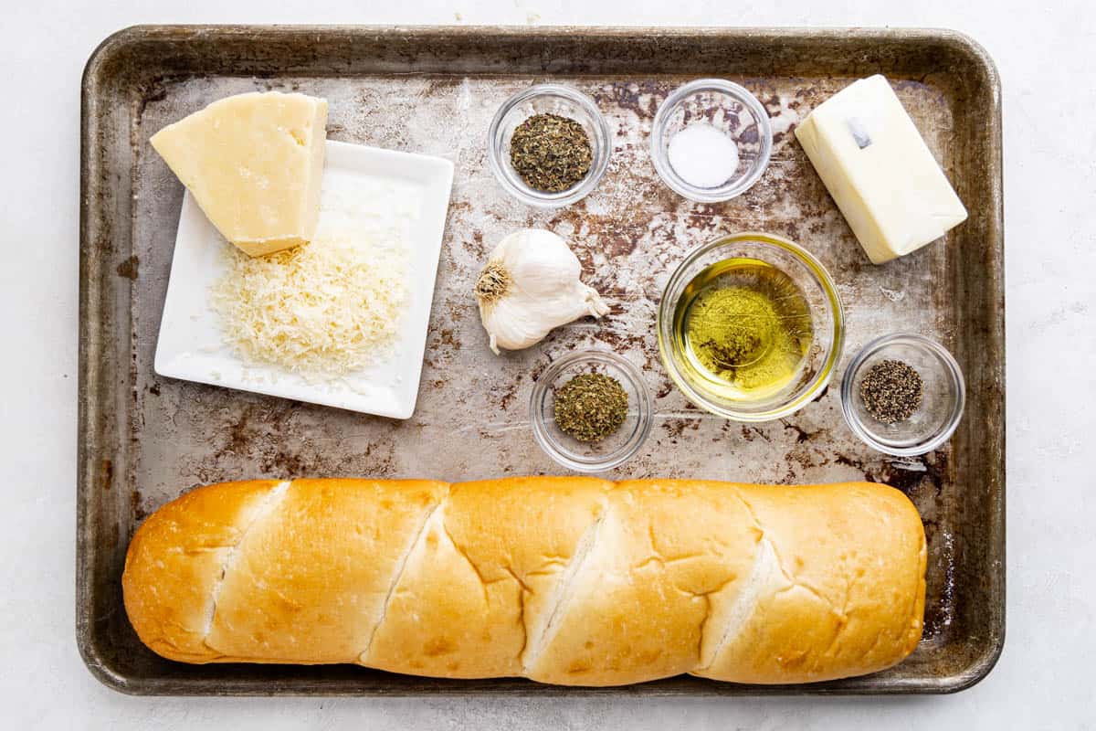 Overhead view of a baking sheet with a loaf of French bread, olive oil, butter, garlic, shredded parmesan, and measured out herbs and spices in glass bowls.