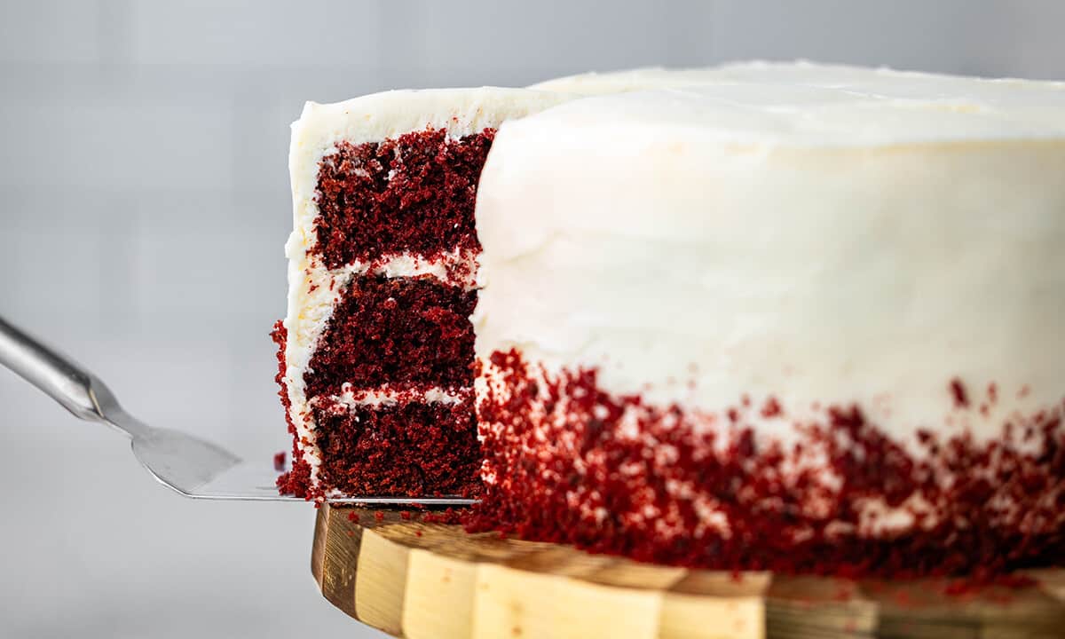 A spatula pulling out a slice of red velvet cake from the whole cake on a gold cake stand.
