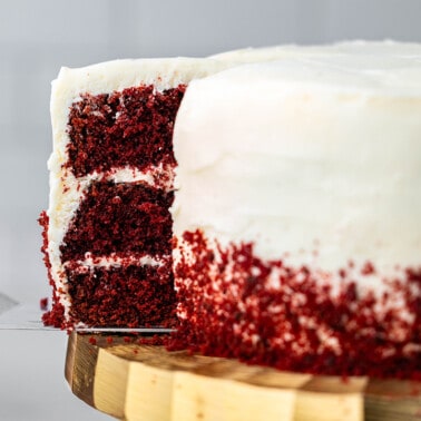 A spatula pulling out a slice of red velvet cake from the whole cake on a gold cake stand.