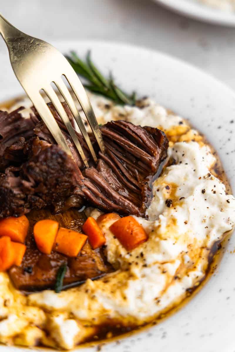 A fork shredding a braised beef short rib served over mashed potatoes.