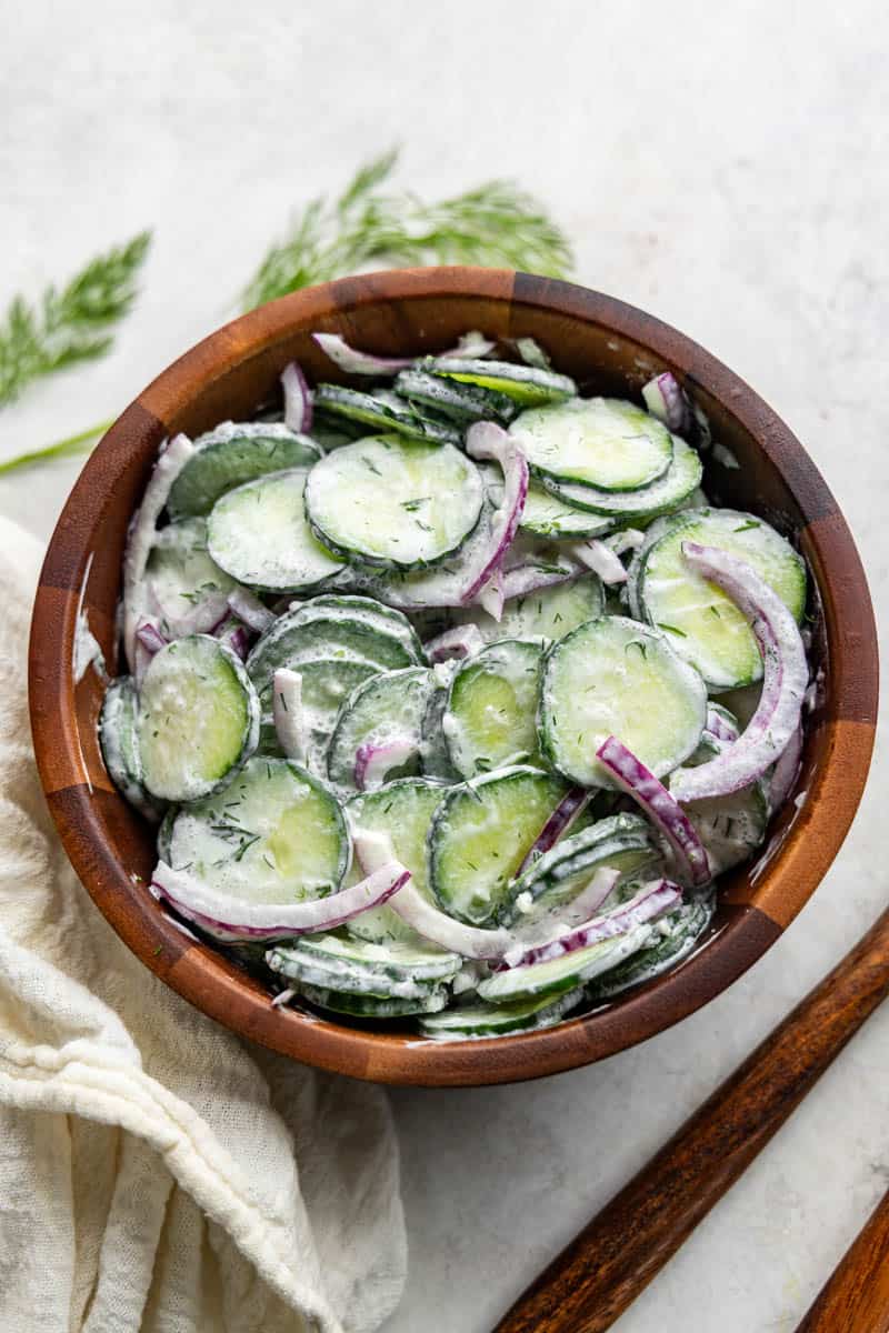 An overhead birds-eye view of a wooden bowl filled with creamy cucumber salad with wooden serving utensils on the side.