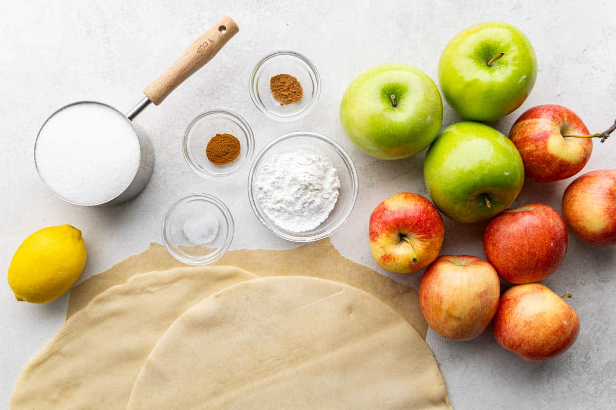 Overhead view of a kitchen counter with pie dough, sugar, apples, flour, lemon, and spices.