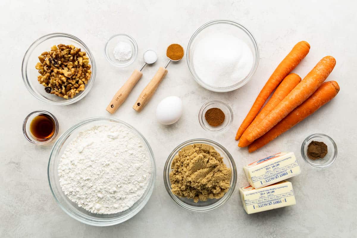 Overhead view of a kitchen counter with measured out walnuts, flour, sugar, egg, butter, brown sugar, carrots, and spices.