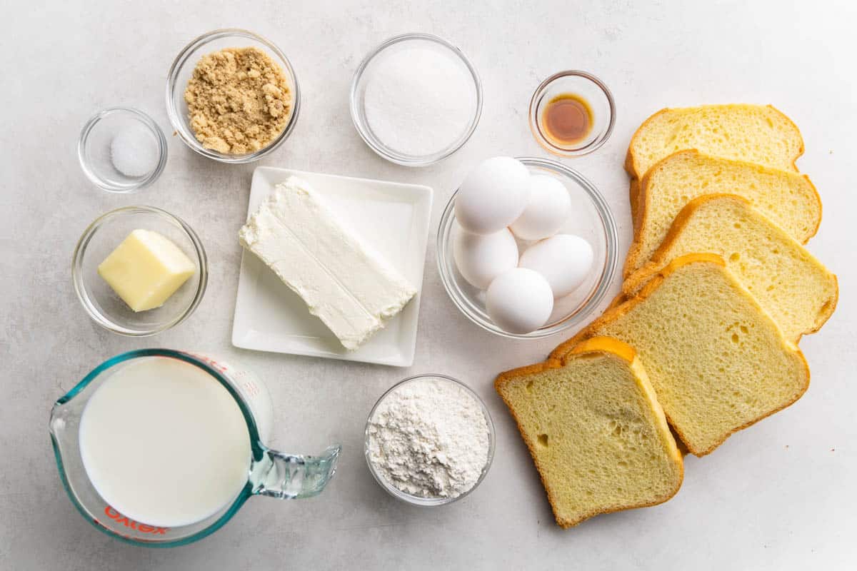 Overhead view of measured eggs, milk, butter, flour, cream cheese, salt, sugar, vanilla, and slices of bread.