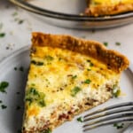 A close up view of a slice of quiche on a plate with the rest of the quiche in the background.