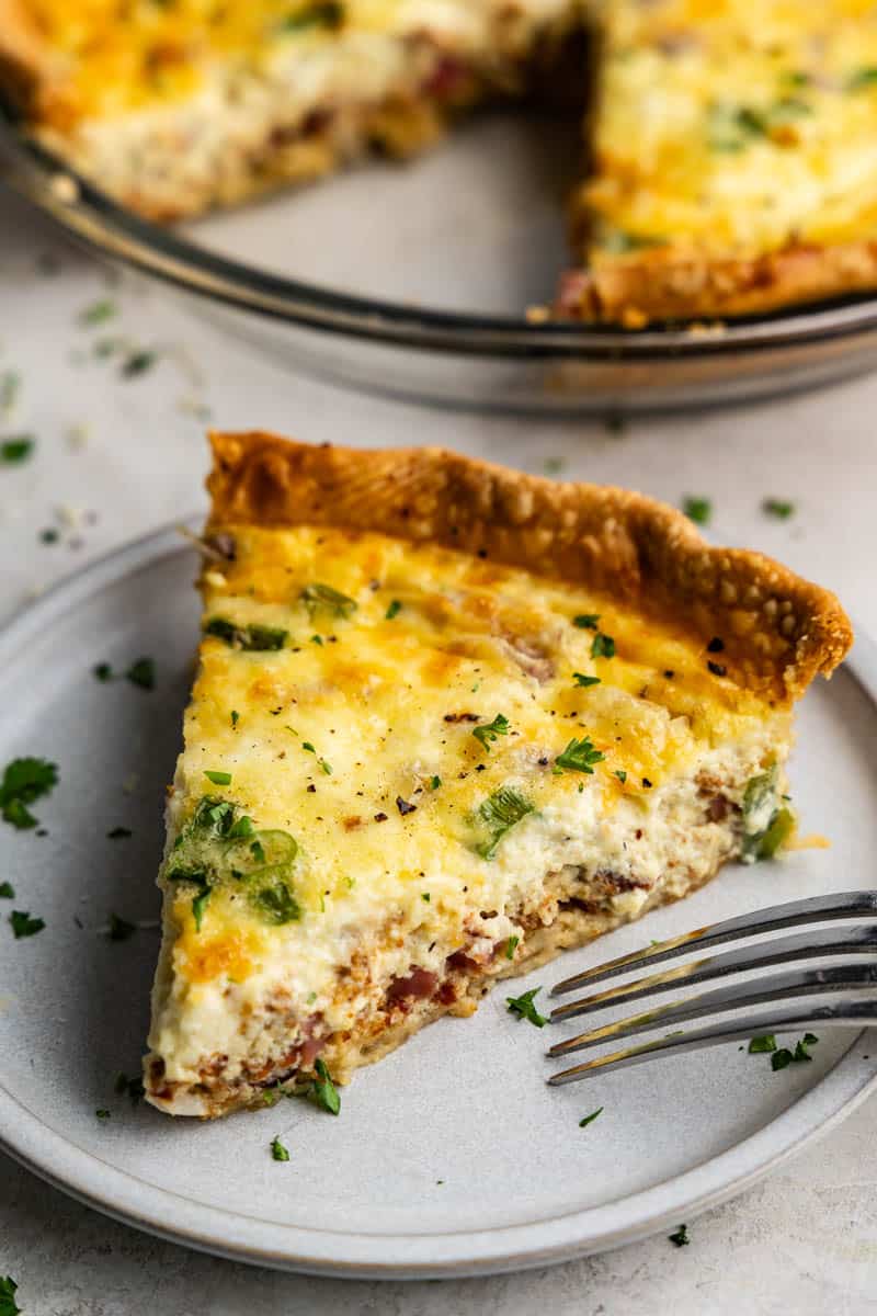 A close up view of a slice of quiche on a plate with the rest of the quiche in the background.