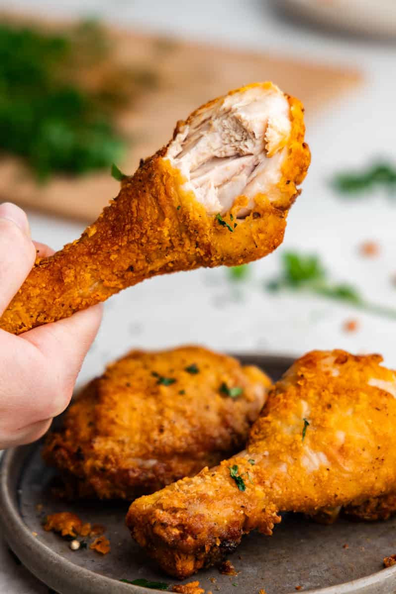A hand holding up a fried chicken drumstick that has a bite taken out of it.