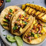 3 tacos al pastor on a plate with grilled pineapple spears on the side.