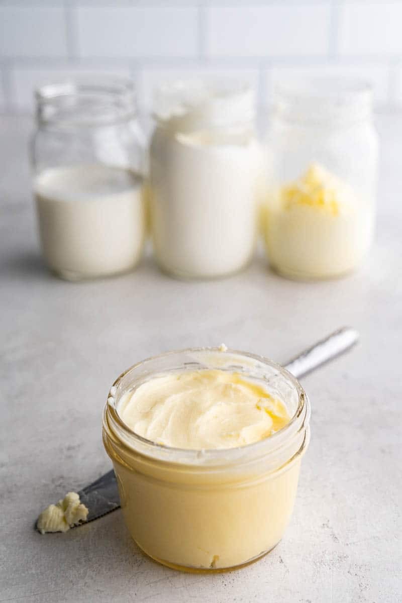 A jar of homemade butter sitting on a counter with 3 mason jars in the background in three different stages of making homemade butter.