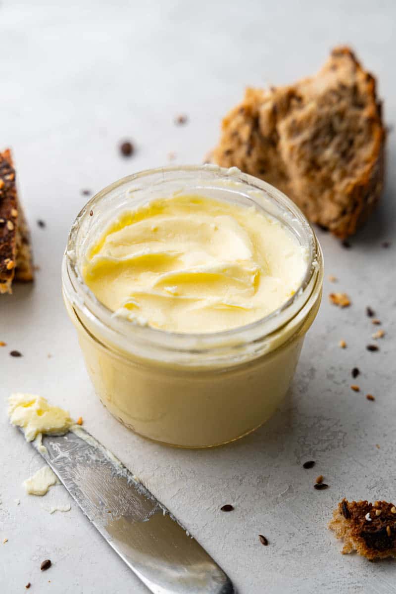 A jar of homemade butter sitting on a counter with chunks of bread in the background.