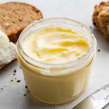 A close up view of a jar of homemade butter with pieces of bread in the background.
