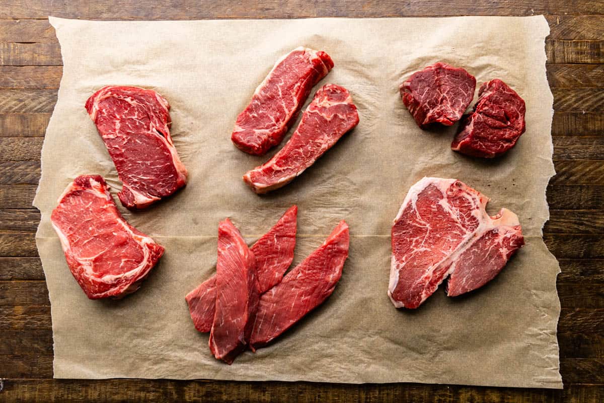 An overhead view of the 5 most common cuts of steak, all raw on parchment paper. 