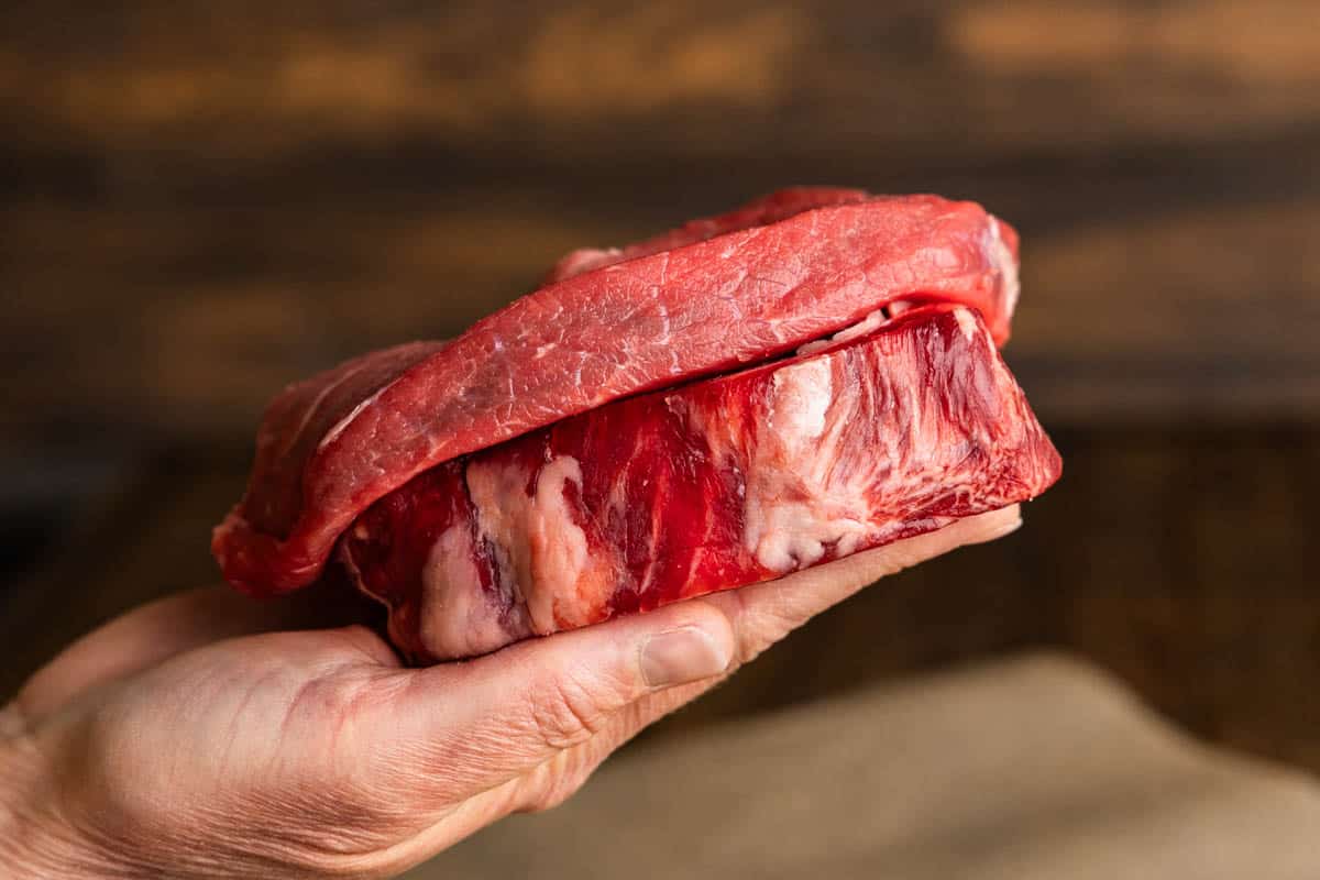 A hand holding two different steaks of different thicknesses. 