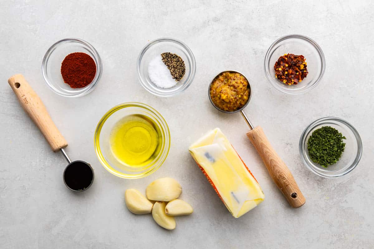 Bird's-eye view image of the ingredients needed to make Cowboy butter. It includes butter, olive oil, garlic cloves, stone-ground mustard, Worcestershire sauce, smoked paprika, dried parsley, salt, crushed red pepper flakes, and black pepper.