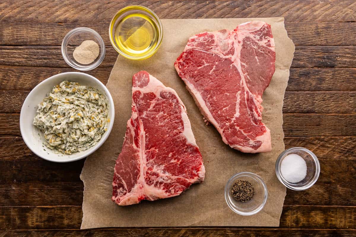 An overhead view of the ingredients needed to cook a t-bone steak including the meat, resting butter, salt, pepper, and oil.