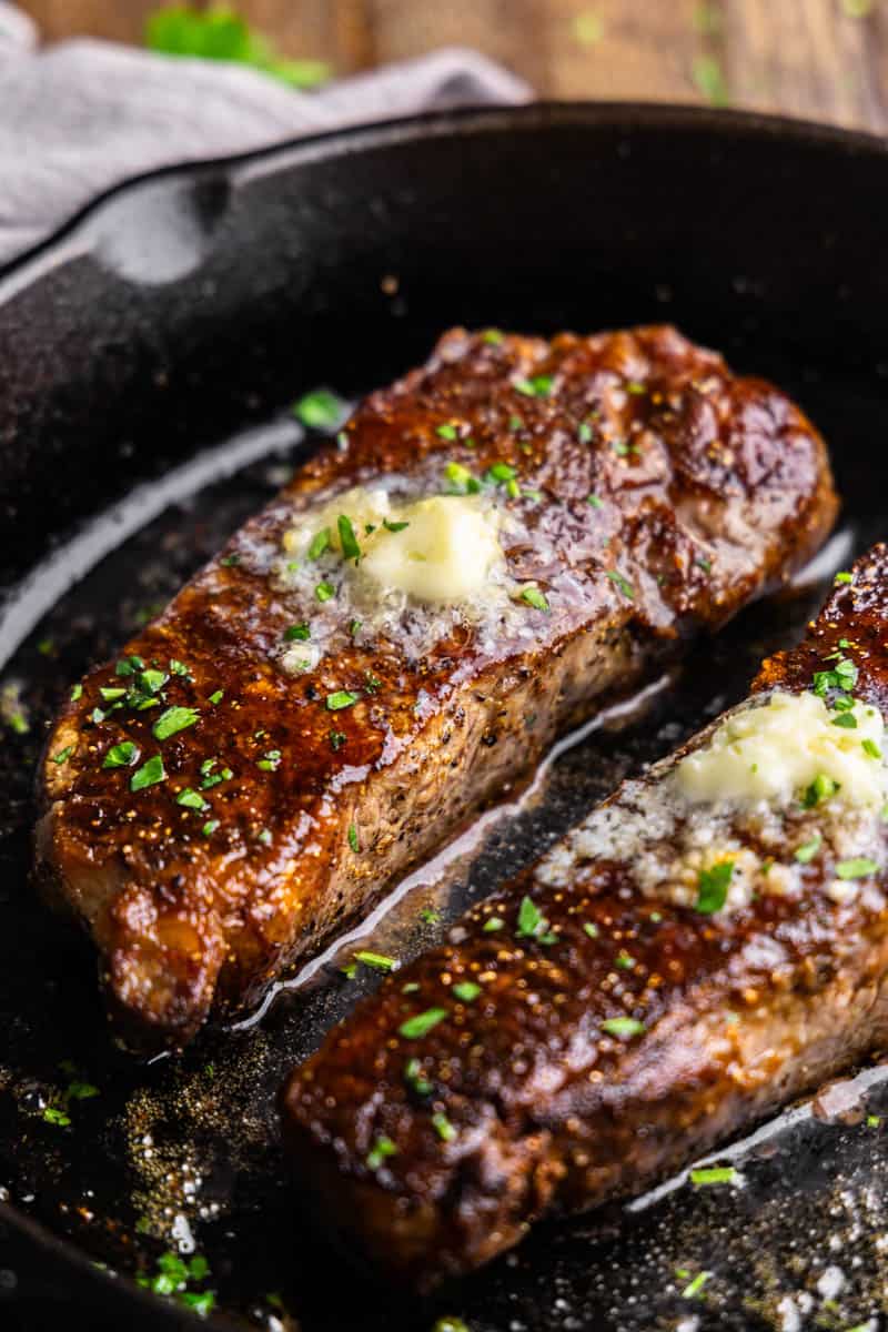 A close up view of two new york strip steaks in a cast iron skillet topped with a resting compound butter. 