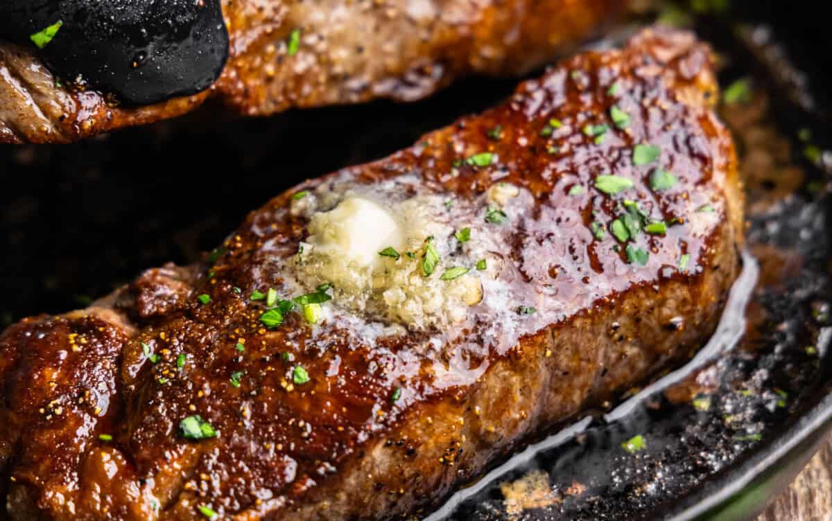 A close up view of two new york strip steaks in a skillet with resting butter on top.