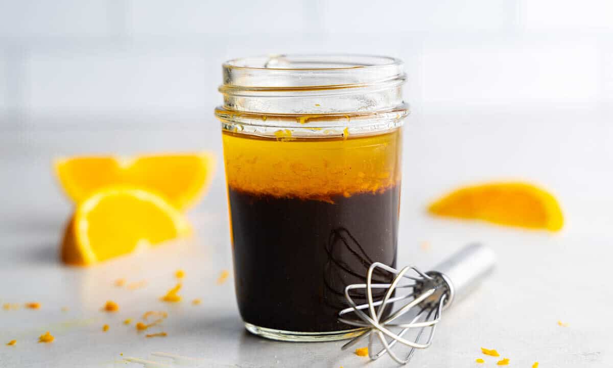 A jar of orange balsamic vinaigrette salad dressing on a table with a whisk and orange slices around it.