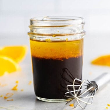 A jar of orange balsamic vinaigrette salad dressing on a table with a whisk and orange slices around it.