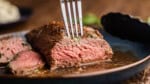 A close up view of a petite tender steak that has already been cut into showing a perfectly pink interior with a fork poking in the top.