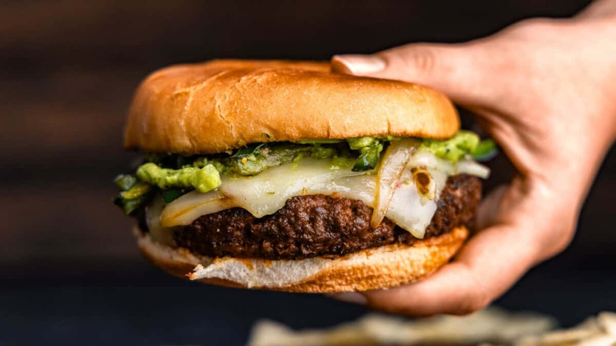 A hand holding a juicy queso fundido burger with cheese and guacamole. 