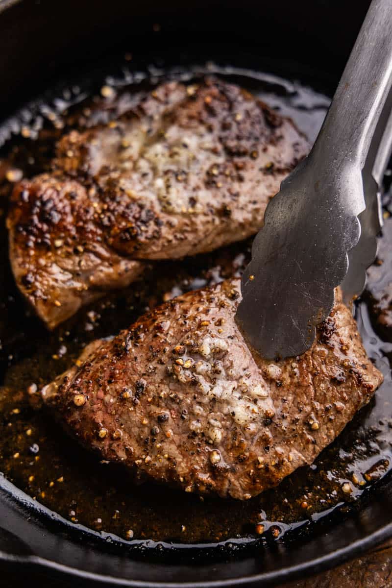 An overhead view of two sirloin steaks in a cast iron pan with metal tongs picking up one steak.