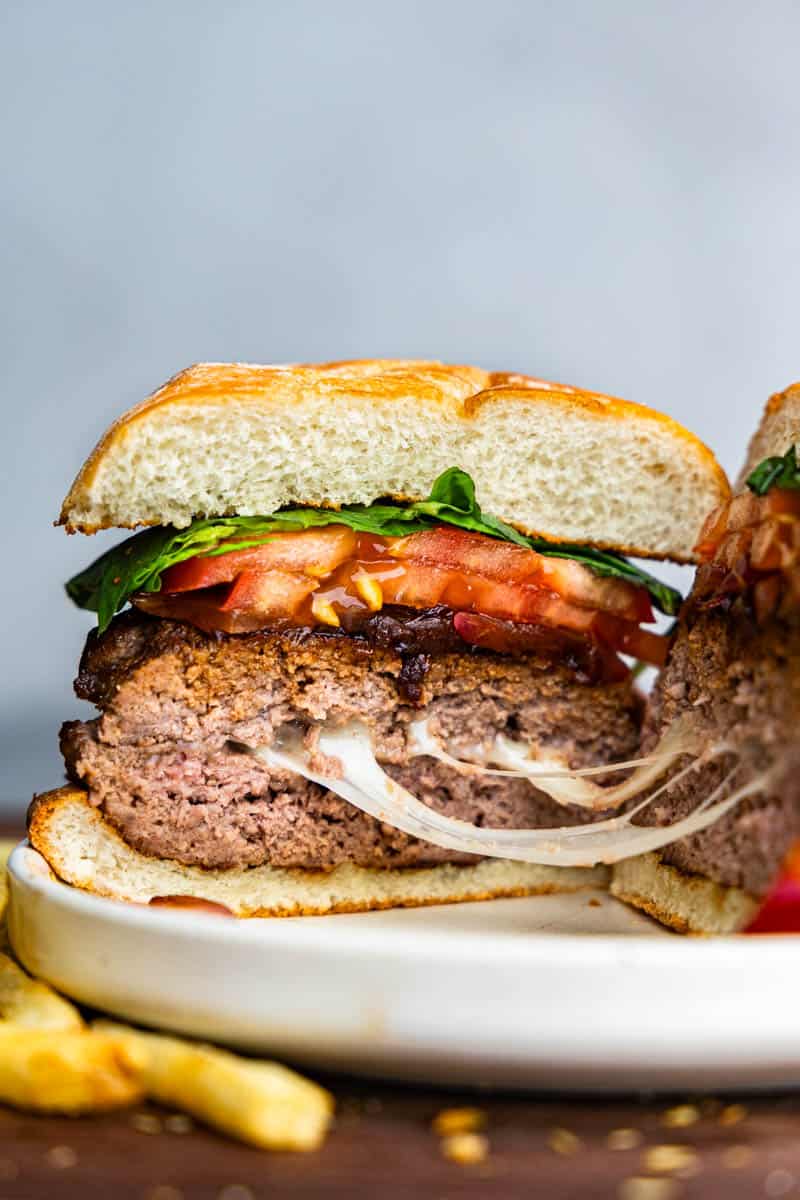 A caprese burger that has been cut in half to reveal strings of gooey melted mozzarella cheese. The burger is topped with balsamic onions, sliced tomato, and fresh basil leaves.