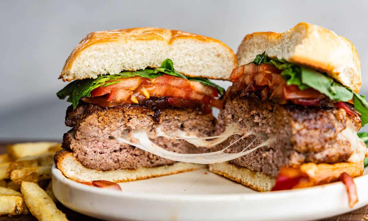 A caprese burger that has been cut in half to reveal strings of gooey melted mozzarella cheese. The burger is topped with balsamic onions, sliced tomato, and fresh basil leaves.