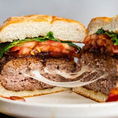 A caprese burger that has been cut in half to reveal strings of gooey melted mozzarella cheese. The burger is topped with balsamic onions, sliced tomato, and fresh basil leaves.