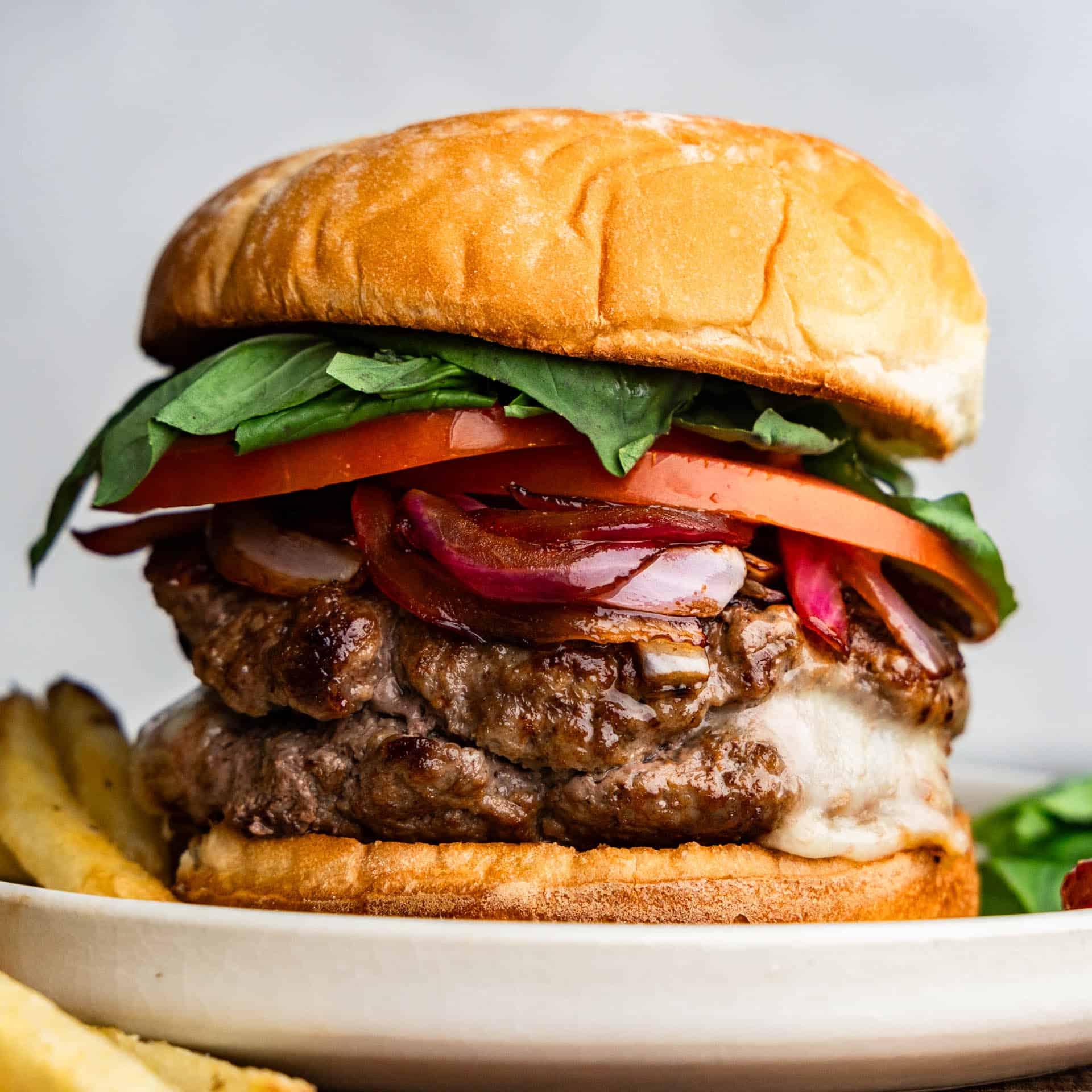 A caprese burger topped with balsamic onions, sliced tomato, and fresh basil leaves. You can see mozzarella cheese oozing out the side.